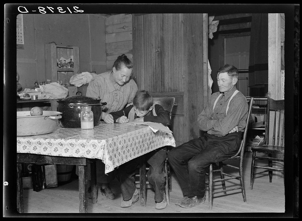 This black and white photograph depicts a family moment in an early 20th-century rural setting. On the left, we see Dora, aged around ten years old, sitting at a table covered with a patterned cloth. She is intently copying out notes from a recipe book on lined paper while her mother stands behind her. The woman appears focused as she pours milk into a jug next to an empty pot; both items rest upon the family's wooden counter which holds various cooking utensils and containers, suggesting they are preparing or have just completed a meal.

Sitting at the table is Dora’s father, dressed in suspenders over trousers with a button-up shirt. He gazes down towards her work on the paper but maintains an observant posture that suggests he may be overseeing his daughter's task while also engaging somewhat with another family member off-camera to his right. His expression seems reflective.

The scene is domestic and filled with quiet activity, indicative of daily life in a working-class rural household at that time period. The wooden cabinetry, simple furniture, and the overall setup are characteristic of modest living conditions typical for families engaged primarily in subsistence farming during such times.

In terms of background context provided by external sources (which is not always available with stock images), this photograph appears to be from a series documenting rural li [...]