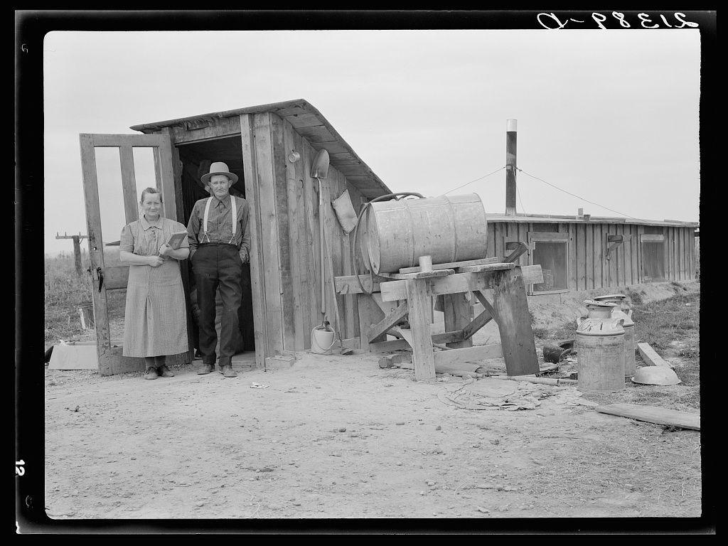 The image is a black and white photograph depicting two individuals standing at the entrance to what appears to be a rudimentary wooden structure, possibly their home. The man on the left wears suspenders over his trousers and has a hat; he stands with one hand in the doorway while holding some papers or documents in the other. Beside him is another person wearing a long-sleeved shirt and skirt. Various items are scattered around them: a large barrel, buckets, rags, and what seems to be household containers. The setting suggests an outdoor rural environment, possibly from early 20th-century America given its style of clothing and the simplicity of construction materials used for housing in such conditions.