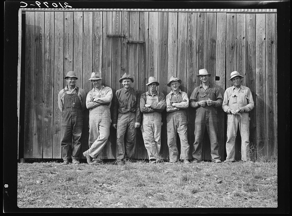 The image is a black and white photograph depicting seven men standing side by side in front of an old wooden barn. They are dressed casually, with most wearing overalls or jeans paired with shirts and hats, indicative of rural work attire from the early to mid-20th century. The men display various poses: some have their arms crossed, others lean slightly against each other, while a few stand confidently upright. Their facial expressions range from serious to smiling.

The setting appears rustic, suggesting an agricultural or farming environment typical of the American Midwest during that era. A small rectangular opening in one panel on the barn suggests maintenance work has been done previously, and there's a visible gap between some panels indicating weathering over time.

Based on textual information overlaid at the top left corner with "D-PROFS," it appears this photo could be part of an agricultural or cooperative project. The text may refer to a name associated with these individuals or their work environment. There is no clear indication of branding, but rather, the image captures what seems like candid workers possibly on break from farming activities.

The photograph has visible wear and tear around the edges, hinting at its age as well as frequent handling over time which indicates it may have historical significance for agricultural heritage studies or rural li [...]