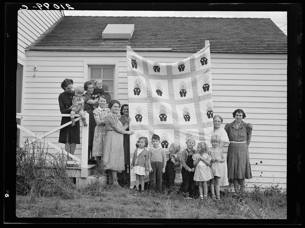 The image shows a group of people, including women and children, gathered outside a house. They are displaying a quilt that features various patterns with images such as cups and saucers or other household items. The individuals appear to be part of the "Helping Hand" club in Yamhill County, Oregon, participating in community service by creating quilts for someone who needs assistance.