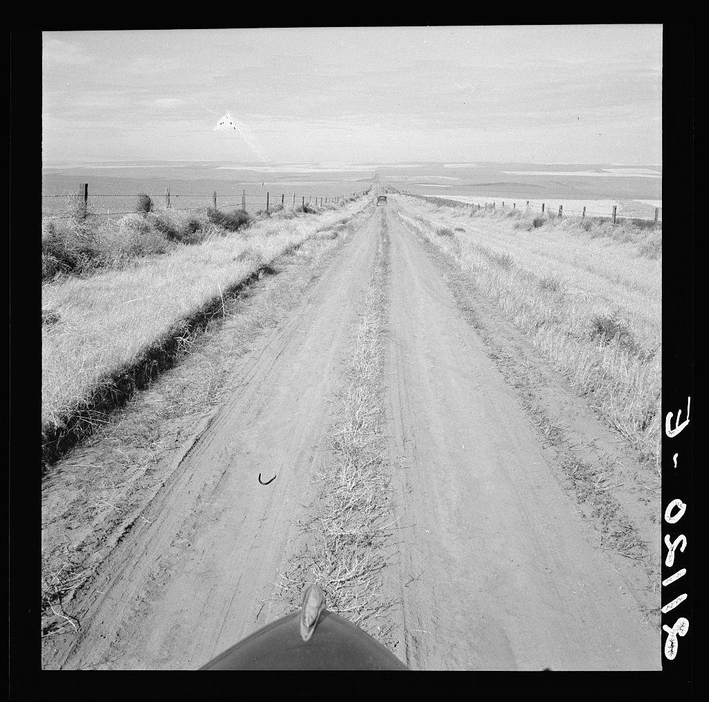 The image shows a black and white photograph taken from the perspective of someone traveling on a rural dirt road. The road stretches straight ahead into the horizon, with fields or prairies visible in both directions as far as the eye can see. On either side of the road are fences supported by wooden posts, which seem to demarcate property boundaries but also allow for some vegetation and shrubs to grow through them.

The sky is overcast, suggesting a cloudy day without much sunlight illuminating the scene directly. In the distance along one stretch of land on both sides, there appears to be a body of water or wetland area with patches reflecting light differently than the surrounding grasses and dirt. The overall environment gives off an impression of aridness and sparseness typical of rural agricultural regions.

The photograph's composition emphasizes solitude and desolation, capturing the vast open space characteristic of certain farming areas in Western states such as Oregon within the United States' western wheat country known for high productivity per acre according to the provided context. This area is also linked with ownership by banks, mortgage companies, and insurance firms which hints at socio-economic implications tied to agricultural land management practices.

The photo bears a copyright mark "Dorothea Lange" in its upper right corner and has an alphanume [...]