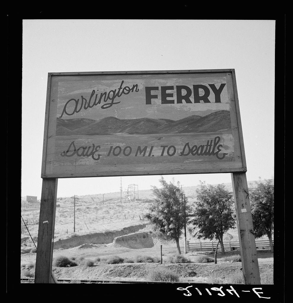 The image is a black and white photograph depicting an outdoor scene. It features a large, vertical sign with the words "Arlington FERRY" at the top in bold letters, followed by smaller text that reads "Drive 100 MI. TO Seattle." Below this main message, there's additional cursive-style writing stating "South & Southwesterly to Arlington," suggesting directions towards the ferry and destination.

The sign is mounted on a wooden post, which appears weathered with age, set against an open landscape that has sparse vegetation including shrubs and what seems like grassy areas. In the background, there's a hilly terrain leading up to electric power lines stretching into the distance, hinting at some industrial or residential structures further away.

The photograph captures this sign in sharp focus while keeping its surroundings somewhat blurred, emphasizing the ferry route information for travelers passing by on Route 97 north of Arlington. The overall setting appears desolate and remote with an arid climate judging from the landscape features visible within the frame.

In terms of context, based on the provided link to a website image titled "On transportation outskirts of small Oregon town," it suggests that this ferry service could be part of rural transport in the state of Oregon. Given Arlington is located near the Columbia River and west-central Gilliam County, such fer [...]