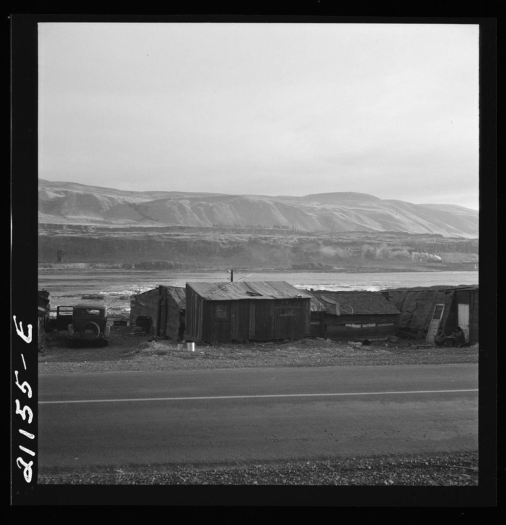 The image depicts a desolate rural landscape with dilapidated structures scattered across the barren ground. In the foreground, there is an old road that stretches into the distance, dividing the frame diagonally and creating a sense of depth. Alongside this road are various buildings, some appearing to be in disrepair or abandoned, characterized by their weathered wood and metal roofs.

Behind these makeshift shelters, a hilly terrain rises sharply towards more mountainous regions with sparse vegetation. The hills seem barren and mostly devoid of trees, suggesting arid conditions possibly influenced by the nearby water body mentioned as part of Yakima Indian Valley on the Columbia River.

The river itself is visible in the midground, its surface calm but reflective, extending across a considerable width before disappearing into the distance near the base of the mountains. A few plumes of smoke can be seen rising from what appears to be industrial structures or machinery along one side of the waterway, hinting at some form of human activity.

Overall, this black-and-white photograph evokes a somber and bleak atmosphere, capturing an isolated community living in challenging circumstances with little sign of modern amenities.