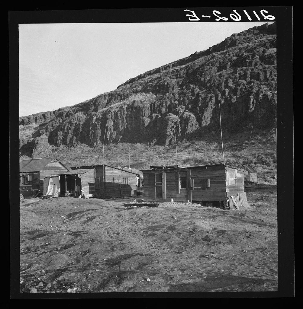 The image depicts a rural scene with several small wooden structures resembling shacks or makeshift shelters. These buildings are scattered across an uneven, rocky terrain that appears dry and barren. In the background stands a steep hillside composed of rugged rock formations extending into the distance under a clear sky.

To provide context to this black-and-white photograph: it captures a snapshot from Yakima Indian village located on the Columbia River in Oregon during salmon season when indigenous people inhabited these temporary shelters for fishing activities. After their time by the river, they returned to live and farm in the valley known as Yakima Valley. This particular photo highlights an incident where locals voiced disapproval of efforts towards improving living conditions through government intervention.

The image bears a label "3-2016," which could indicate its date or perhaps some other reference information related to its documentation within archives like those held by Dorothea Lange, who was known for her work with the Farm Security Administration. The website credit points toward the source being loener.nl and provides further details on this specific photograph's provenance.

The overall mood of the picture is somber; it reflects a lifestyle characterized by hardship but also one that retains cultural traditions amidst challenging environmental cond [...]