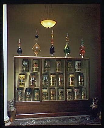 The image displays a collection of glass bottles and jars, each with different colored labels and shapes. They are neatly organized on shelves within a wooden cabinet that has multiple compartments. The cabinets appear to be placed against a wall in an indoor setting. There is a small hanging light fixture above the display case. In the background, there seems to be a partial view of another object which looks like a vase or urn with flowers near its base.
The image appears to have historical significance as it was taken at an American Institute Pharmacy and dates approximately from 1920-50s based on the context provided in "American Institute of Pharmacy. Jar and bottle display at American Institute of Pharmacy II ca. 1920-ca. 1950." The photographer is credited to Horydczak, who was active during that time period.
The image contains a single transparency with dimensions approximately 8 x 10 inches as specified in the source information provided: "1 transparency : color ; 8 x 10 in" from https://images.loener.nl/horydczak/full/691e/691ec6a0450e57a2674cf539.jpg.