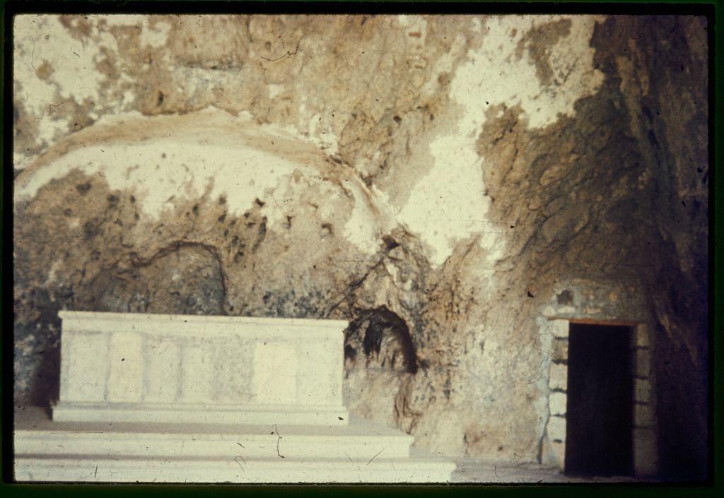 This image depicts an ancient, weathered cave with rough stone walls. The focus is on a large white sarcophagus or tombstone situated at the forefront of the frame. This marble structure appears aged and eroded in places due to natural elements and time.

In the background, there's an entrance to what seems like another chamber within the cave system, characterized by its dark interior which contrasts with the lighter tones around it. The setting gives off a historical or archaeological ambiance, suggesting that this might be a site of religious or cultural significance, possibly connected to St. Peter as mentioned in Galatians 2:11.

The overall tone is earthy and muted, dominated by browns and whites from the stone surfaces, with splashes of color highlighting certain areas such as the dark doorway on the right-hand side of the image. The photograph has a vintage feel due to its sepia-like hues and slight graininess, indicative that it could be between 1950 and 1960.

The caption "Along the Mediterranean coast, southward" suggests geographical context while mentioning Antioch hints at religious or historical significance associated with St. Peter's shrine within a cave setting as per Galatians' verse mentioned in the additional information section provided alongside this image.