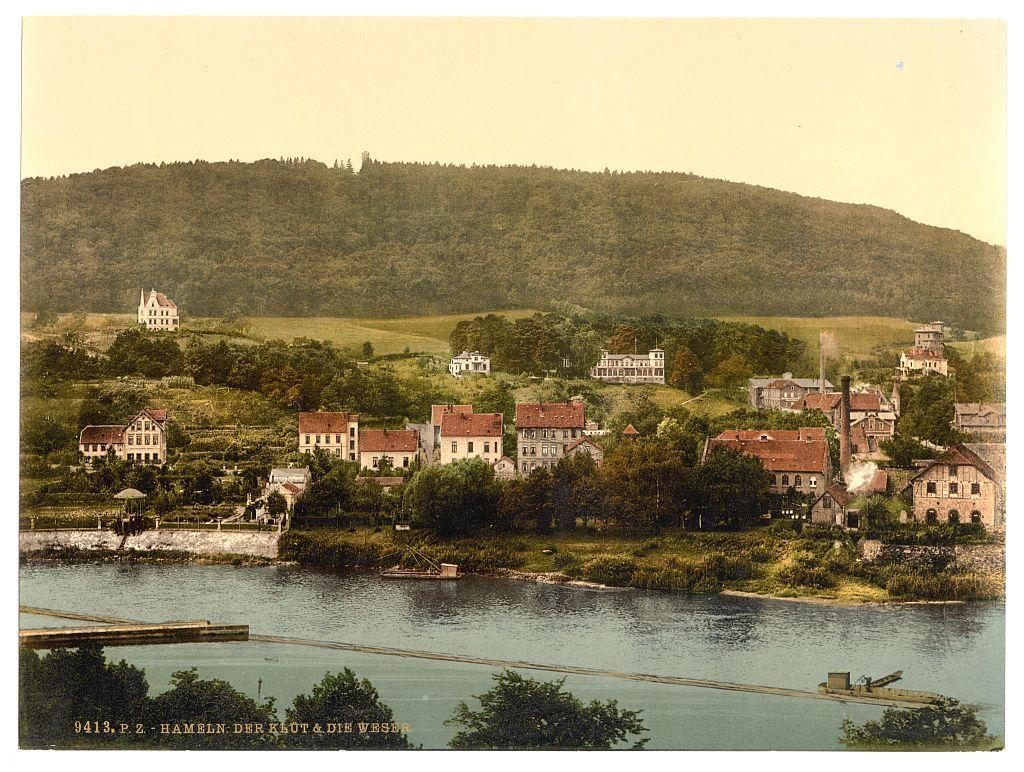 This image depicts a historical scene, likely from the late 19th to early 20th century. The photograph shows an idyllic village nestled on elevated ground overlooking a river with visible structures like houses and buildings featuring red-tiled roofs. In the background is a lush green hill or small mountain range that appears dense in vegetation. A body of water, possibly part of the Weser River based on the caption "Der Klut & Die Weser," flows through the foreground alongside some sort of man-made structure resembling an aqueduct or bridge for boats. The overall color tone has faded to a sepia hue, suggesting it's either aged significantly or processed intentionally to give it that effect.

Additional information about this image indicates its origin and historical context:
- "Der Klut & Die Weser" refers to locations in Hameln (Hanover), Germany.
- It is dated between ca. 1890 and ca. 1900, placing the photograph within a period of industrialization or modern development when such scenic views were often captured for postcards or prints.

The image captures an early representation of German rural life with architectural styles characteristic of that era and geographical features typical to Northern Germany's landscape.