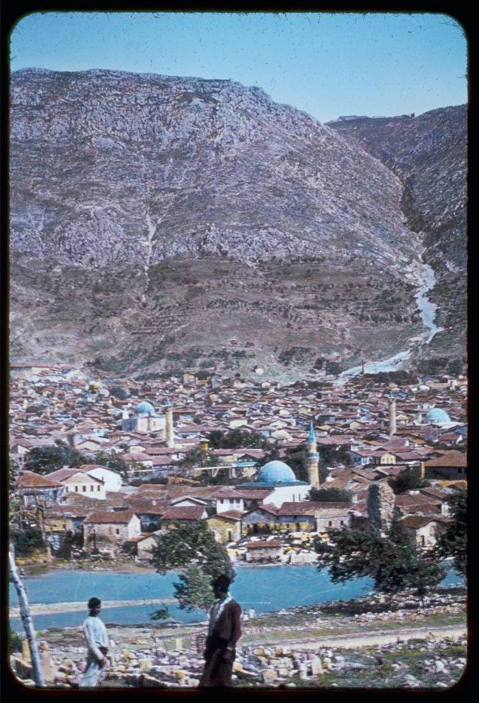 The image depicts a picturesque village nestled at the foot of rocky, hilly terrain. The town features an array of buildings with terracotta roofs and several prominent blue-domed mosques that suggest Islamic architecture, indicative of this area's cultural heritage. A river or stream meanders through the landscape towards what appears to be a larger body of water in the foreground, possibly part of a lake system.

There are two individuals visible at the bottom left corner; one is walking while another seems to pause for a moment. The person closer to the camera wears light-colored clothing and has their head down, looking as if focused on something out of view. Another figure stands further away in darker attire near what appears to be a waterfront area with piles of goods or materials.

The overall scene conveys an everyday glimpse into life within this historical community.