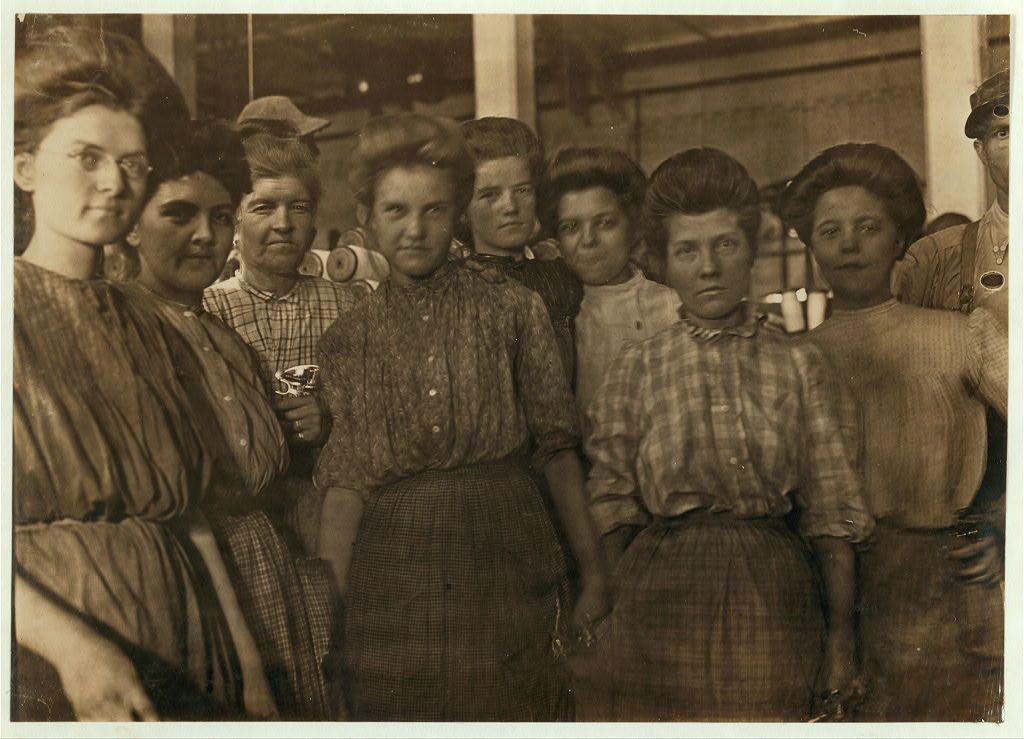 The image is a historical black-and-white photograph showing ten individuals who appear to be women from the early 1900s. They are dressed in working-class attire, with many wearing buttoned-up blouses and skirts or dresses of that era's style. Their hairstyles also reflect late Victorian-era trends: tightly pinned updos common among female factory workers during this period.

The group is standing closely together, suggesting they might be colleagues or part of a community. One woman in the center holds what looks like a glass mug or cup with her right hand, while another person on the left side seems to hold an object that could possibly be related to their work environment.

In the background, there appears to be some industrial equipment and materials which may indicate this photo was taken at a factory. The presence of such items suggests these women might have worked in a mill or similar establishment during this period. This image is part of Lewis Hine's collection documenting labor conditions under capitalism for workers like those employed by the Bibb Manufacturing Company, specifically their textile factories located around Macon.

The sepia tone and wear on the photo indicate it could be over one hundred years old, providing a glimpse into historical working-class life in America. The image is labeled with descriptive information suggesting its origin as part o [...]