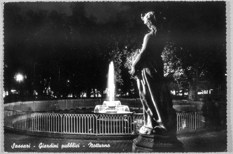 The image is a black and white photograph capturing an evening scene in a public garden. A prominent feature of the photo is a statue, possibly depicting a female figure with flowing hair, standing on what appears to be a pedestal or base adorned with inscriptions that are not legible from this angle. The statue's right hand seems raised slightly above its body level.

In the background, there is a fountain with water spouting upwards; it stands in the center of an enclosed space defined by a metal railing. This enclosure suggests it might be a plaza or central feature within the garden.

The lighting indicates that the photo was taken at night as artificial lights illuminate parts of the scene and create contrasts between light and dark areas, giving depth to the photograph. The darkness surrounding the lit subjects adds to the ambiance, likely enhancing its nighttime setting.

Intriguingly, text appears in two different languages - Italian ("Sassari" which refers to a city) followed by "Giardin publici," suggesting it's an image of Giardini pubblici (public gardens), and then English at the bottom right corner with the word "notturno," meaning night.

The watermark or signature on this photo is "FerroCandilera" which could refer to a collection, photographer, or organization associated with such works. The mention of 'VEDUTA;GIARDINI PUBBLICI' implies that it might be p [...]