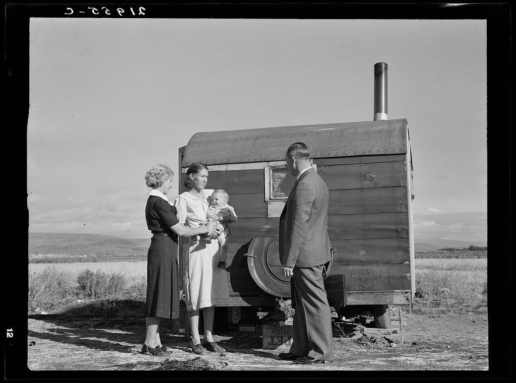 The image captures a candid moment of interaction between three individuals and an older, wooden trailer. Two adults, one in a dark dress with light-colored shoes and the other wearing business attire including trousers and a coat, engage closely with each other while holding a baby dressed in white garments. The setting appears to be outdoors on dry land, likely indicative of rural or agricultural surroundings, given its simplicity and openness.

The trailer, which serves as an informal shelter or living space for these individuals, has visible characteristics like a door and a chimney pipe that suggests it's equipped with basic amenities such as heat. Its construction is utilitarian, reflecting the practical needs rather than comfort. The absence of modern technology hints at a time period possibly linked to mid-20th century rural life.

The photograph is in black-and-white, which emphasizes contrasts between light and shadow, adding depth and texture that might have been lost with color photography during this era. This monochromatic quality also lends the image an air of historical context, potentially placing it within a timeline where such trailers were common as temporary housing solutions for those involved in transient rural work.

The photograph is marked "3-268" at the top right corner and has text that seems to read 'Dorothea Lange', which identifies Dorothea  [...]
