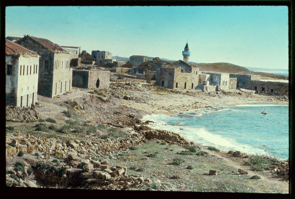 The image depicts a coastal scene with remnants of ancient architecture and ruins. On the left side, there are stone buildings that appear to be in various states of decay, suggesting historical significance or abandonment over time. Among these structures is one building with an ornate dome-like structure on top, possibly indicative of Islamic architectural influence.

In the background, a hill rises gently against a clear sky, and further back, another tower with a distinctive blue-tinted spire stands out prominently. The middle ground shows more stone ruins scattered across uneven terrain leading down to a rocky shoreline where waves lap against the beach.

To the right is an expansive view of calm sea water that meets the horizon under what appears to be bright sunlight. There's also a lone sailboat on the far side, suggesting recreational or small-scale fishing activities in this tranquil setting. The landscape includes patches of vegetation and dry earth with scattered rocks, giving it a rugged feel.

The photograph is labeled as part of an archive from Matson Photo Service, which indicates that these images might have been used for travel documentation or historical research purposes during the 1950s to mid-70s.