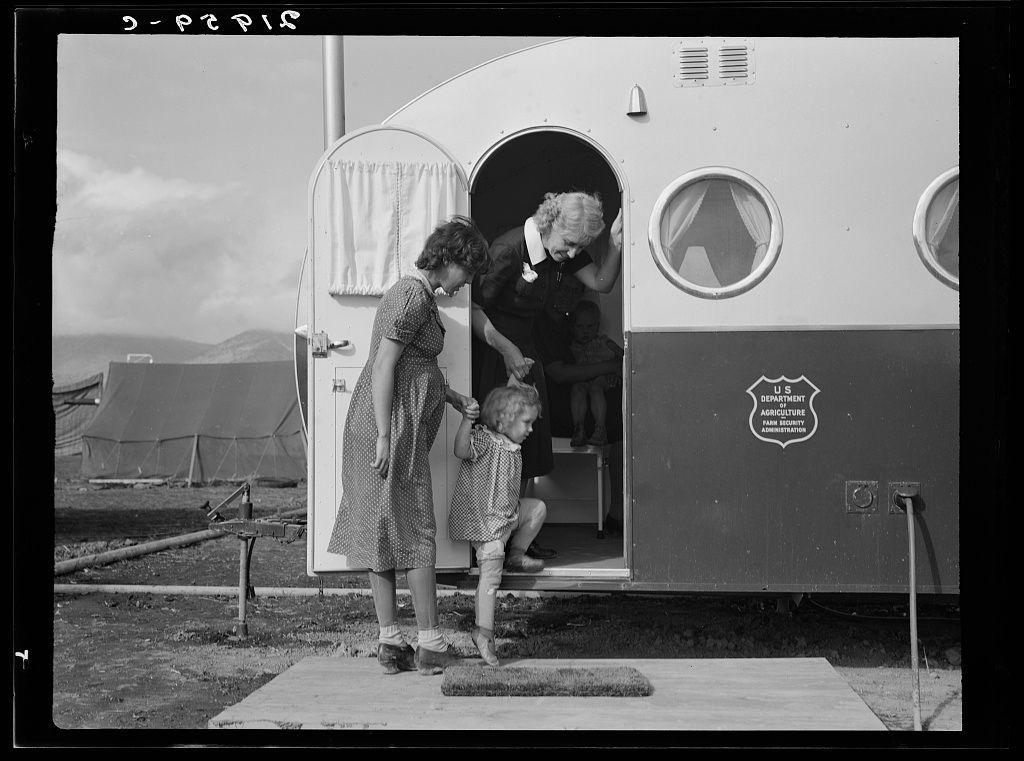 The image depicts a scene from what appears to be an era with mid-20th-century fashion, featuring three individuals near the entrance of a trailer. A woman is holding onto the handrail and stepping out of or into the trailer, which has curtains on its windows and doors, characteristic of portable housing units typical for traveling purposes during that time period.

Inside the trailer, another person is seated with their legs visible above what seems to be medical examination tables. The interior looks simple but functional, suggesting a temporary setup possibly used in remote locations or events requiring mobile services such as health examinations.

Outside, on the ground near where these individuals are situated, there's an item resembling a mat for stepping onto before entering and another similar object close by which could serve as support when getting up from sitting. A child stands beside the woman who is exiting the trailer; they appear to be dressed in casual attire of that period.

The setting seems outdoor with tents visible in the background indicating this scene might take place at a camp or temporary settlement, possibly related to agricultural activities given the "U.S. DEPARTMENT OF AGRICULTURE" sign on the side of the mobile unit, which suggests it could be connected to rural assistance programs like those by FSA (Farm Security Administration).

There is [...]