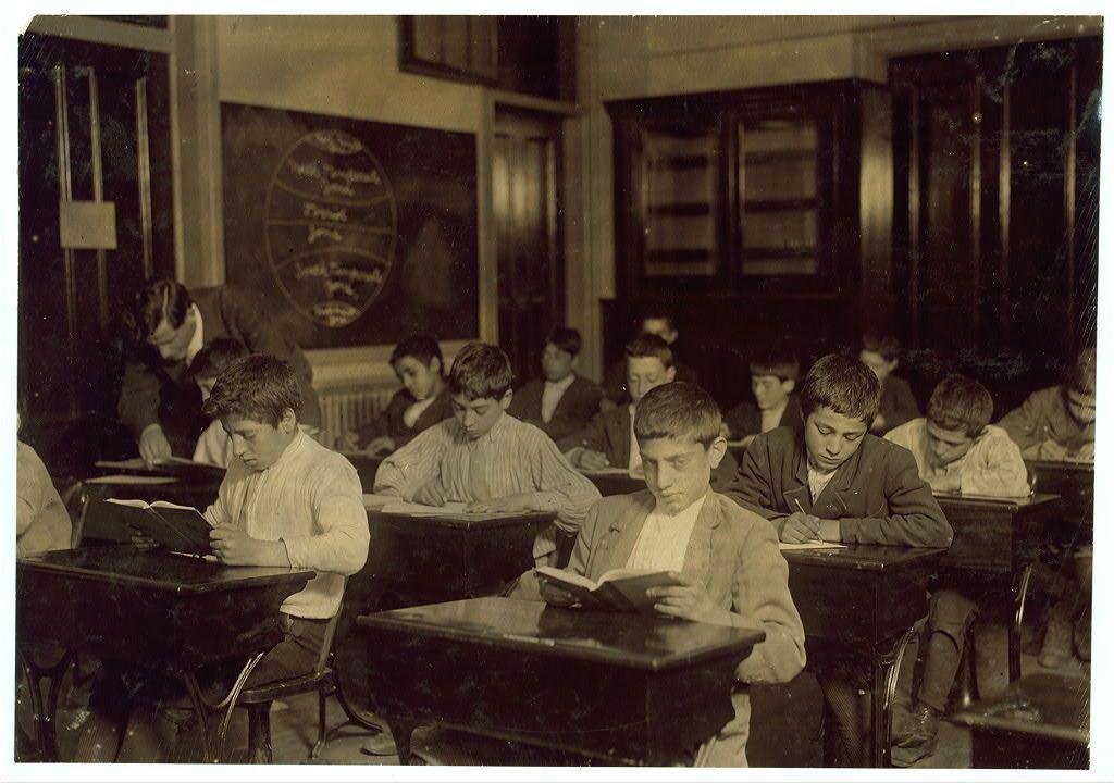 The black and white photograph captures a group of students seated in an old-fashioned classroom, focused on their studies. They are using desks with metal frames and wooden surfaces typical of the early 1900s. The room is furnished with dark wood cabinets filled with books or documents along one wall, and there's a large chalkboard at the front displaying what appears to be lesson plans or class schedules written in cursive script. Some students are engrossed in reading from open textbooks while others write notes on small pieces of paper placed directly beside their desks.

In terms of attire, they wear period-specific clothing such as long-sleeved shirts and jackets for boys, with no visible accessories that would indicate a modern setting or time frame beyond the late 19th to early 20th century. The overall ambiance suggests an educational environment dedicated to learning from written materials in a structured manner.

The photograph is credited to Lewis Hine through the provided link which indicates it comes from "Immigrants in night school" and specifies its location as Boston, Massachusetts. This context adds historical depth to the image, highlighting not only the academic setting but also potentially reflecting educational practices or immigrant community involvement during that era.