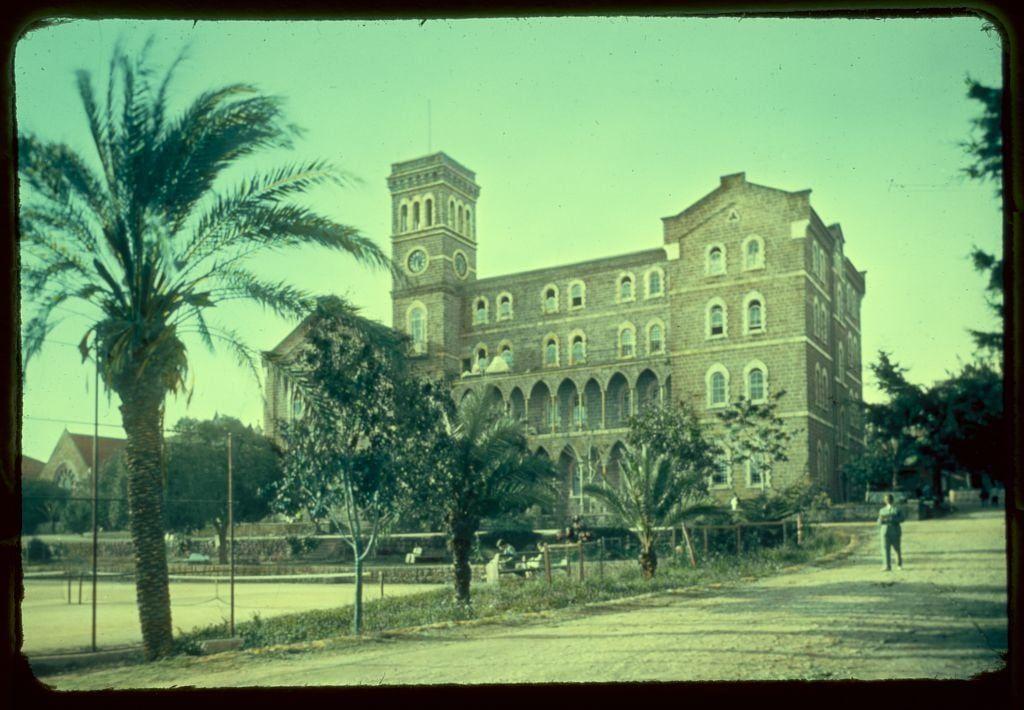 The image depicts a historical building with European architectural influences, possibly in the Mediterranean region. The structure is multi-storied and features arched windows on its facade, typical of such buildings often used for institutional purposes like schools or hospitals during that era.

In the foreground, there's a palm tree and some greenery indicative of a temperate climate. A paved road curves through the bottom part of the picture with a person visible in motion on it, suggesting pedestrian activity near this building. The sky is clear with no apparent clouds, hinting at sunny weather conditions during the time when this photo was taken.

This photograph appears to be from an archival collection related to Beirut's College Hall between 1950 and 1977, capturing a moment in its history before it underwent any significant alterations or events.