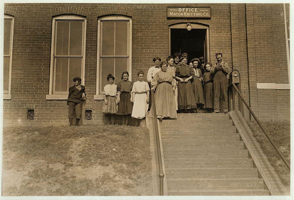 The image depicts a historical scene with an assortment of individuals standing at the entrance and on steps leading up to what appears to be an office door labeled "Macon Knitting Co." This photo seems to have been taken in Macon, Georgia during early 20th-century work hours. The people include men, women, and children dressed in clothing consistent with that era's typical attire for various occupations. Some wear long-sleeved shirts or blouses while others are clad in dark overalls suitable for manual labor. Their poses range from standing upright to resting against the building, suggesting a casual gathering during their workday break time known as "noon hour." The architecture of the brick structure includes large windows with security bars and a modest entryway leading into an office or administrative area.