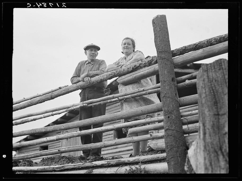 The image is a black and white photograph depicting two individuals, presumably a man and woman, leaning on the horizontal rails of a wooden fence that appears to be part of an old log cabin. The structure suggests rustic simplicity typical of early frontier living or rural life in certain regions during the time period it was taken.

This particular photo seems to capture a moment from 1935 as indicated by the date '2-48'15 printed on the negative, which is part of its border. It's likely associated with an agricultural setting given the individuals’ clothing and posture that exudes laborious undertakings common in rural or farm communities.

The man wears a cap, long-sleeved shirt, trousers, and sturdy shoes suitable for manual work. The woman, dressed modestly, appears to be wearing a dress typical of early 20th-century attire with her hair pulled back into a practical style. Her arms are crossed over the fence, while his hands rest on its railing.

The setting is an outdoor scene possibly in Idaho as referenced by the provided caption hinting at rural development or agricultural activity during that period and location. The image's content evokes themes of hardship, resilience, family life, and community endeavor prevalent among settlers during times like the Great Depression when such conditions were common for many Americans.

Given additional context from a larger  [...]