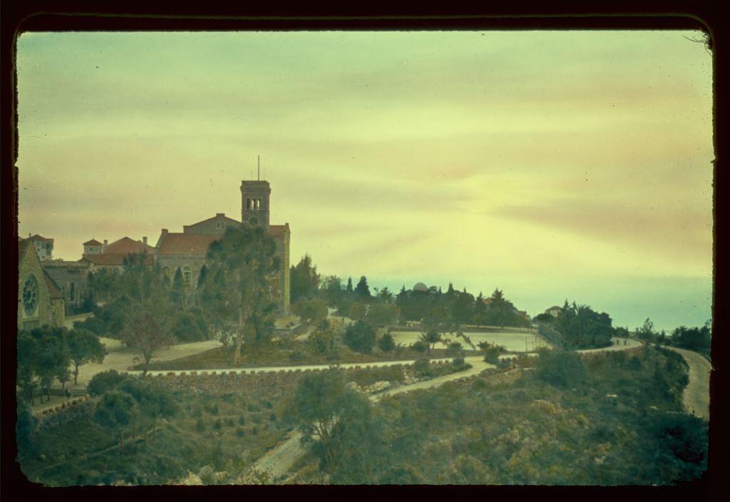 The image depicts a picturesque scene of an architectural structure, likely situated in the Mediterranean region given its proximity to Beirut. The focal point is a grand building with Gothic elements such as pointed arches and large windows, which could be identified as part of the American University of Beirut campus during 1950-77 period based on historical context provided. This university stands prominently within an expansive landscape that transitions into hilly terrain covered in greenery.

The photograph captures a tranquil setting with trees gently swaying alongside winding roads leading up to and around this prominent building, suggesting it is perched atop a hill or elevated area for visibility purposes possibly tied to its institutional significance. The sky above displays a serene ambiance characterized by soft pastel hues of yellow and orange near the horizon gradually blending into lighter shades as they reach towards the clear skies.

In summary, the image portrays an idyllic view of an educational institution's campus nestled within natural surroundings, set against the backdrop of a calm yet vibrant sunset.