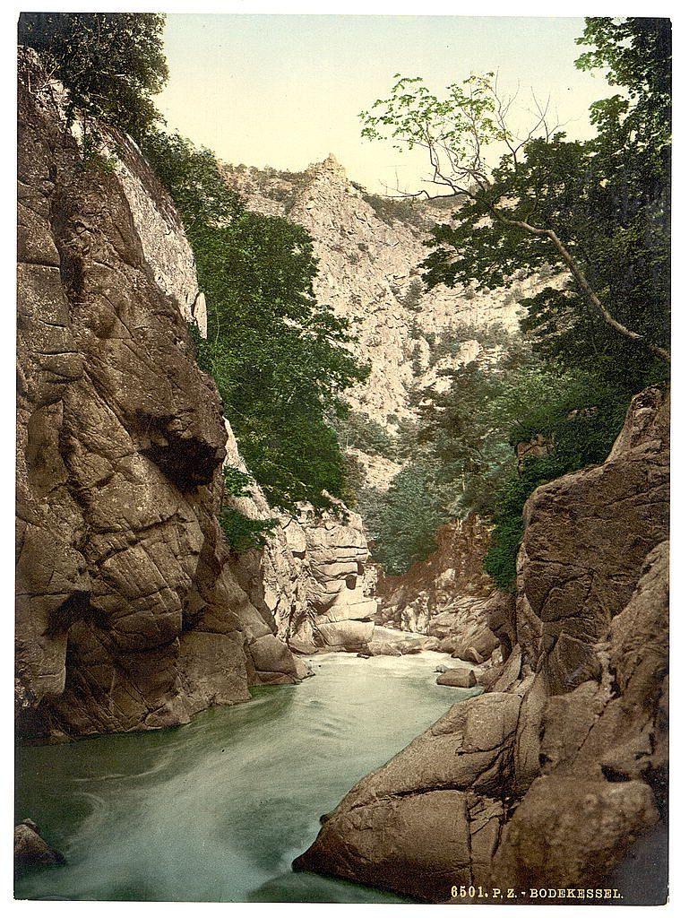 This image depicts a natural landscape featuring a river flowing through a rocky canyon or gorge. The water appears calm and reflects the colors of its surroundings, suggesting that it may be shallow in some areas near rocks protruding above the surface.

The rock formations on either side are rugged with varying textures and shades ranging from light beige to brownish tones. Green vegetation is visible among the crevices and along the sides of the canyon, hinting at a temperate climate where such flora can thrive.

In the background, there's an elevation or hill that extends into what appears to be a misty atmosphere, indicating it could be part of higher terrain like mountains or hills. The sky is not clearly visible but seems overcast based on the diffused lighting in the image.

At the bottom right corner, there's text "6501.P.Z.- BODEKESSEL," which may refer to a catalog number for this photograph and possibly indicates that it was taken by a photographer or someone named Boëdkessel. This suggests historical context as such watermarking is common in old photographs.

The overall impression is of an untouched, serene environment with natural beauty emphasized by the presence of water amidst stony surroundings. The scene seems to be captured during daylight hours when there's enough light to highlight details within the canyon but not so bright that it washes out featu [...]