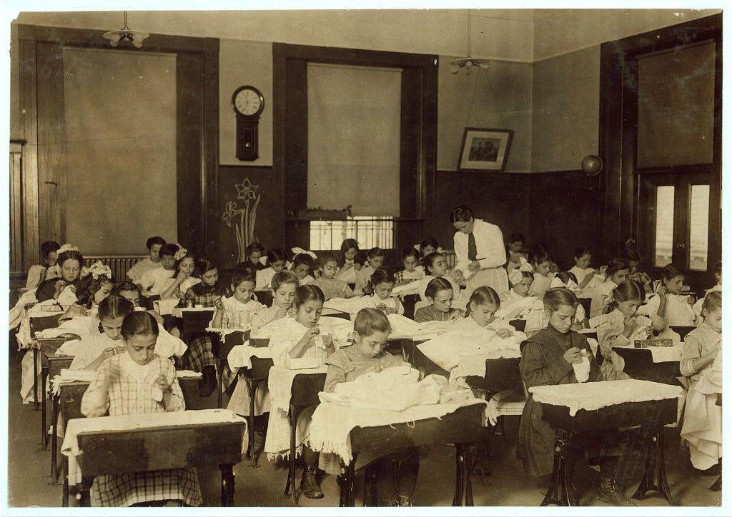 The image depicts a classroom from the early 20th century, filled with students seated at desks and engaged in various activities. The room is furnished with dark wood benches and chairs for each student, arranged neatly rows. Each desk appears to have an attached shelf above it where books or materials may be stored.

In this setting, children are focused on tasks such as writing, reading from books, or working on cloths laid out before them, possibly part of a domestic science class activity given the context suggested by their clothing and workstations draped with white linens. The students are dressed in traditional early 1900s school uniforms for girls that include knee-length dresses often with plaid skirts.

A teacher is standing at the back near a large clock on the wall, overseeing the classroom's activities. Additional decorations like floral designs add character to the room. Sunlight seems to be coming from windows out of frame or artificial light sources as indicated by the overall lighting conditions within the space. There are also items such as globes and possibly other educational tools hung against the walls.

The photograph has a vintage, monochromatic feel indicative of early photographic technology. It is labeled with text indicating it may have been taken in Boston, Massachusetts during 1915 at Hancock School for pupils identified as "the steamer Class."