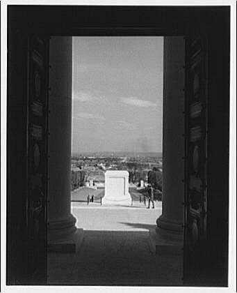 The image is a black and white photograph taken from the perspective of someone standing inside an open doorway. Through this opening, we can see two large stone columns framing either side of what appears to be an outdoor area with several people walking around.
In the middle ground between the pillars lies a prominent monument, seemingly made of marble or another light-colored stone material. The structure is rectangular in shape and features smooth surfaces on all sides. Its design suggests it could be a memorial or tombstone.
The overall setting has an open sky above, indicating that this location likely serves as some sort of outdoor amphitheater or gathering space overlooking the surrounding area. A few individuals can be spotted at various distances within this expanse: one person walking away from us to our right and another closer but facing in a different direction towards what could possibly be trees.
The photo seems historical, suggesting it was captured during an earlier era based on its black-and-white composition as well as the attire of those present.