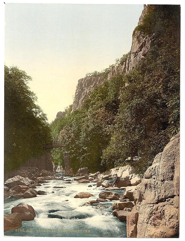 This image depicts a picturesque natural scene, featuring a river flowing through a rocky canyon with lush greenery on either side. The sky is overcast with soft lighting conditions suggesting it could be early morning or late afternoon. A wooden bridge spans across the river at an elevated position in the middle of the composition. Large boulders and rocks are scattered along the water's edge, adding to the rugged beauty of the landscape. No people or animals can be seen, emphasizing a sense of tranquility and untouched nature. The colors range from deep greens of the foliage to varying shades of blue and white within the moving river waters.
