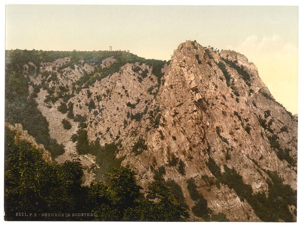 This image depicts a rugged mountain landscape, featuring steep cliffs with rocky surfaces and sparse vegetation. The terrain is uneven and appears challenging to navigate. A small section of the cliff has structures that resemble human-made pathways or trails carved into its surface. In the distance on top of the highest peak stands what looks like an observation tower or monument, suggesting a place of interest for visitors or locals. Trees with green foliage are interspersed throughout the lower slopes and around the base of the mountain, providing contrast to the rocky terrain. The sky is clear with a soft gradient from light yellow near the horizon to pale blue above, indicating either early morning or late afternoon sunlight. At the bottom left corner, there's text "6521-P.Z.- SOHURHEIM IM BODETHAL," which seems like an identification marker for this particular photograph within Schurre in Bodethal, Hartz region of Germany between approximately 1890 and 1900.