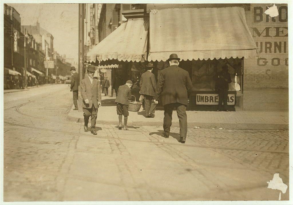 The photo is a black and white image that captures an urban street scene from the early 1900s. The perspective shows pedestrians walking on what appears to be cobblestone streets, indicative of older city infrastructure typical in many historical American cities during this era.

There are at least seven individuals visible; three men who seem engaged with each other and two others possibly conversing or simply passing by. A young child is also present, accompanied closely by an adult male, likely a parent, as they walk down the street together. The adults wear hats that were common for both fashion and protection from sun exposure at this time.

In the background are several commercial establishments with awnings extended over their entrances, suggesting it could be market day or simply everyday commerce in what looks like a busy district of Boston given the caption's reference to Boy Marketing in Boston. Storefronts display items that would have been accessible for purchase by early 20th-century consumers.

The atmosphere is one of daily life and business as usual; however, there’s no sign of modern amenities or technology which places it firmly into a historical period well before digital age advancements were common in everyday society.