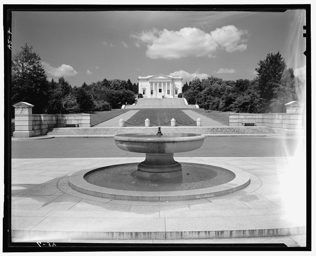 The image is a black and white photograph of the Tomb of the Unknown Soldier at Arlington National Cemetery. The perspective is from a distance, showing a large circular fountain in the foreground with a rectangular water basin on top. Behind it stands a grand neoclassical building flanked by trees under a clear sky. Several stone structures flank either side leading to stairs that ascend towards this impressive monument. It appears to be an open area within Arlington National Cemetery as indicated by surrounding greenery and pathways.