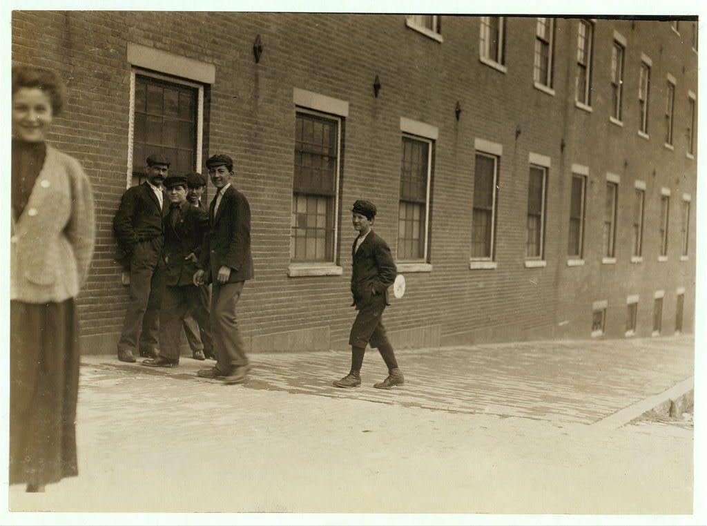 The image depicts a sepia-toned photograph from what appears to be the early 1900s, capturing several individuals in front of an industrial brick building with multiple windows. There are four adults and one child visible; all but the woman on the far left wear hats, which were common for both men and boys at that time as a signifier of social class or occupation.

The man to the right seems older than his companions and appears more formally dressed in a dark coat, vest, trousers, and a flat cap. He is smiling broadly towards the camera. The other three adults are younger; they wear similar attire but without hats, suggesting they might be employed there rather than visiting guests or customers.

The child stands apart from this group, holding what looks like a tin can in his left hand while walking away with purpose, perhaps heading home after work hours. He wears dark trousers and shoes, matching the adults' clothing style of that era.

In front of them is an unpaved street leading to the brick building where they're standing; it shows signs of wear and tear indicative of a working-class environment. The architecture suggests this was likely in an urban setting during industrialization when such buildings were common for factories or mills employing many workers, including children like our young subject.

This image carries historical significance as part of Lewis Hin [...]