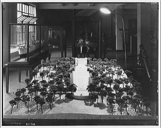 The image shows a model of an approach to the Tomb of the Unknown Soldier, likely from around 1920-1950. It is presented in black and white, suggesting it could be historical or archival material. The scene features a large display with numerous trees lined up symmetrically on either side leading towards what appears to be a monument at the end. A pathway bisects the tree-lined area, converging toward this monument.

The model seems elaborate, depicting details such as individual trees and possibly small structures within them or along the sides of the path. The background includes architectural elements like columns and glass panels, which may indicate an indoor setting meant to showcase the scale and design concept for a real-life space dedicated to honoring an unknown soldier's tomb. A figure can be seen in the far distance at what could represent this monument.

The photo is credited to Horydczak, Theodor (1890-1971), who seems to have captured or created this model display around 16:2A. Additional context for understanding this image and its significance can be found in Arlington National Cemetery archives, which likely includes historical models of various sites within the cemetery grounds.

The overall composition suggests a thoughtful consideration of design elements meant to create an impactful entrance leading up to a revered location, reflecting on themes of re [...]