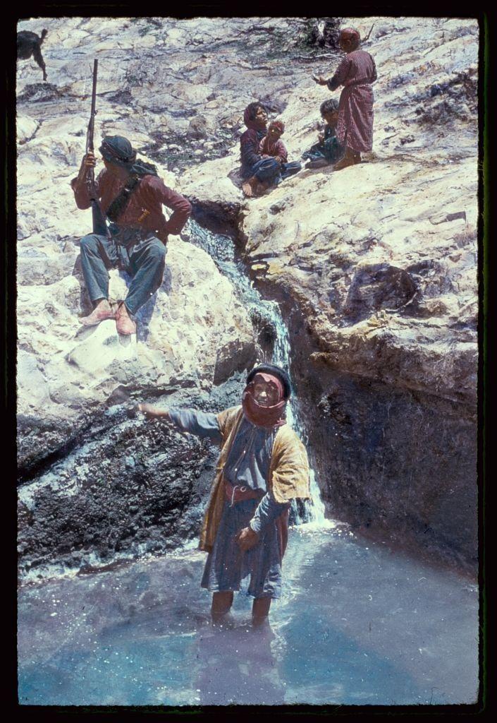 The image depicts a group of people, including both adults and children, situated around a natural water source. In the foreground, an individual stands in ankle-deep water with one leg extended outward towards the camera while extending their arm for balance or support. This person is wearing loose-fitting clothing that appears to be traditional, possibly from a Middle Eastern context. Behind this figure are three other individuals; two of whom sit on rocks and appear engaged in conversation or contemplation, holding what seems like small objects between them.

In the background, another group sits near an open water source, with one person kneeling beside it while others hold onto their belongings. One seated individual is visible from behind wearing a headscarf, suggesting modest dress customs common to certain regions of Middle Eastern descent. The landscape itself consists of rocky terrain which suggests arid conditions typical for such environments.

The colors in the image are muted and naturalistic with earthy tones dominating due to both clothing coloration and the sandy ground surrounding the water source. There's a sense of tranquility despite some individuals being armed, suggesting that this might be part of everyday life within their community during challenging times or circumstances requiring vigilance.