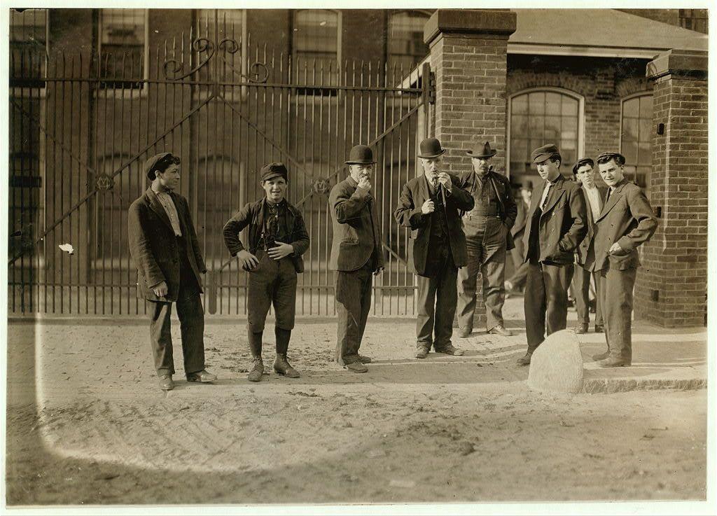 The image depicts a group of nine men standing outside an industrial building during the early 1900s. The setting appears to be in front of Mill No. 1 at Cocheco Mfg. Co., located in Dover, New Hampshire, as noted by the provided caption and reference link. This particular photograph was taken on May 15, 1909, during a specific time of day: 6:00 A.M.
In this black-and-white photo, the men are dressed in work attire from that era, including suits, hats, coats, and overalls. Some wear bowler or derby-style hats, while others have flat caps with ribbed crowns. The group exhibits a mix of ages, ranging possibly from young adults to middle-aged individuals.
One man stands out as he holds up what seems to be an earpiece connected by wires, suggesting that the image might capture workers who had recently been on strike and were waiting for their contracts or payback after returning to work following some form of industrial action. The men are standing in a line with varying expressions; some appear contemplative, while others look more engaged.
The background features a wrought-iron gate partially open behind them, leading into the mill's premises which is not fully visible due to the angle at which this photograph was taken. A brick building and windows can be seen further back, establishing an urban industrial setting typical of that time period in America. The ground appears  [...]