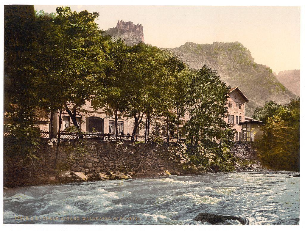 The image depicts a picturesque riverside scene, likely captured in the late 19th or early 20th century based on its style and coloration. A sturdy stone wall borders the water's edge, separating it from a large building with multiple stories featuring numerous windows, balconies, and what appears to be an attic level. The structure is surrounded by lush greenery, including dense trees that frame the scene at various angles. In the background, imposing cliffs rise steeply against a hazy sky, suggesting a mountainous landscape.

The river itself has turbulent waters with visible white foam indicating rapid currents or rapids. A few large rocks are partially submerged in the water close to the bank. The overall atmosphere of the photograph evokes a sense of tranquility and natural beauty, capturing what appears to be an idyllic setting for leisurely activities such as boating or sightseeing.

Additional information about this image can be found on "Views of Germany" website (https://images.loener.nl/PhotochromPrints/full/65e4/65e47a825890ae069f67ec20.jpg). This source provides further details and context for the photograph, indicating its origin from Thale in Bodethal, Germany. The image is also associated with Waldkater (Thale) and was created between approximately 1890 to around 1900, placing it within a time frame that would have been during or shortly after German expan [...]
