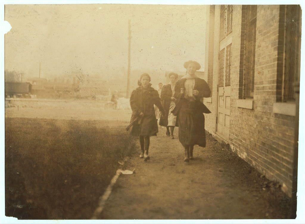 The image is a sepia-toned photograph showing several children walking on what appears to be an unpaved path beside a brick building. The setting seems rural or semi-industrial, with buildings and bare trees in the background suggesting it might be late fall or winter.

In the foreground, three girls are prominently featured: one running ahead with her head turned towards the camera, another following closely behind, both wearing dark coats suitable for cold weather; a third girl is walking beside them, also dressed warmly. Their attire includes hats and scarves, indicative of an era when children wore such garments.

The image carries historical significance as it depicts child labor from approximately 1908 to the early years of World War I in Moberly, Missouri at Brown Shoe Factory, where Eva Robinson was employed possibly around eleven or twelve. The photo captures a moment of their daily commute between work and home life during that tumultuous time period.