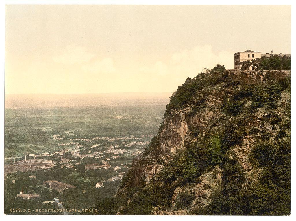 The image is a vintage postcard depicting an elevated perspective overlooking the cityscape and surrounding landscape of Hexentanzplatz & Thale in Germany. The foreground shows rugged, rocky terrain with sparse vegetation indicative of mountainous or hilly regions. A large building sits atop this elevation on the right side, suggesting it could be an observation point or a significant structure within the town.

Beneath lies the urban expanse stretching out towards what appears to be flatland, possibly agricultural fields and plains beyond the immediate vicinity of Hexentanzplatz & Thale. The architecture is dense with structures that suggest residential buildings interspersed among industrial facilities like factories. This mix indicates a small-town setting likely engaged in various forms of economic activities including agriculture and manufacturing.

The sky above has an overcast, muted ambiance suggesting either early morning or late afternoon light conditions. Overall, the image captures a historical snapshot of this German town's layout during its postcard era, showcasing both natural scenery and urban development from a commanding vantage point on elevated terrain.