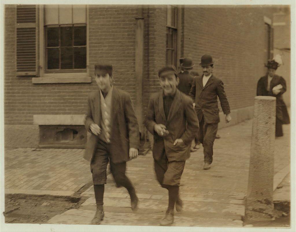 The photograph shows a group of men walking on an urban street during what appears to be the late 19th or early 20th century. The sepia tone indicates that it is likely taken in black and white, as color photography was not widely available at this time. In the foreground are three individuals; two appear younger with a playful demeanor, while one has an older appearance marked by a mustache and bowler hat.

The young men are dressed in work-appropriate clothing of the era: suits, hats, coats, and sturdy shoes. Their expressions range from smiles to laughter as they stride forward on a cobblestone path beside a brick building with windows that have shutters. One man is slightly ahead, leading the others down what seems like an incline.

In the background, another figure wearing similar attire walks in the opposite direction, suggesting this could be part of a work routine or community event within Lawrence, Massachusetts at Pacific Mills during a time when mills were significant employers for many local workers. The presence of multiple individuals and their collective activity indicates a sense of daily life captured candidly without posing.

This photo is linked to Lewis Hine's works on labor conditions in American industries, specifically focusing on the textile mill environment where young boys often worked long hours as part of child labor practices that were common  [...]