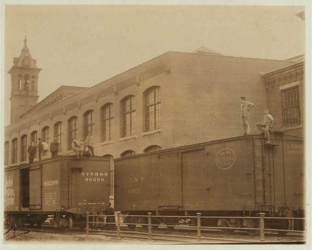 The image depicts a sepia-toned photograph featuring an urban scene with individuals engaging in activities atop and around large railcars labeled "Nynh 1" on the side, suggesting they are part of a freight train. The setting appears to be industrial or warehouse-like buildings with arched windows, indicative of older architectural styles commonly found near railway yards.

Several men can be seen: some perched precariously at the edge of railcars while others stand atop them, possibly waiting for their turn to ride down; one individual is on a bicycle. The individuals are dressed in work attire typical of late 19th or early 20th-century industrial workers. A prominent clock tower looms over the background, adding an element of historical significance.

The overall atmosphere suggests downtime during working hours—typically known as 'dangerous noon-hour'—a time when such recreational activities were common among factory employees seeking brief respite from their labor-intensive jobs. The image is part of a collection that highlights moments in American industrial history and captures the spirit of workers amidst their challenging conditions.