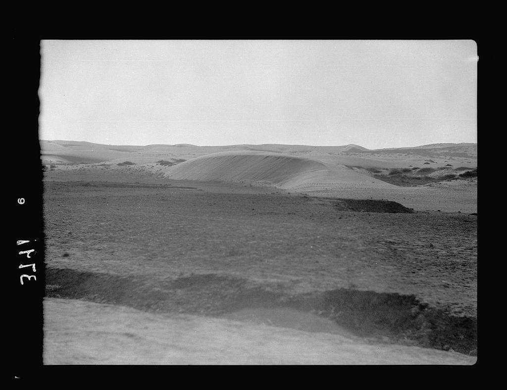 The image depicts a black and white photograph of an arid landscape, likely taken during the period between 1920 to 1933. The scene shows rolling hills formed by shifting sand dunes that are encroaching on nearby farmland along the Mediterranean coast. These natural features suggest dry environmental conditions typical for certain regions around this time and place.

The photograph appears aged, with a visible crease down one side, indicating it may have been part of an album or bound collection. The image has some text at the bottom left corner reading "THE 6," which could be indicative of its cataloging within that group, but without further context on how this was organized, more details cannot be provided.

The American Colony in Jerusalem is mentioned as a possible source for this photograph, hinting that it may have been taken by or associated with the organization. The Photo Department referenced here likely refers to an archival collection of photographs documenting life and landscapes during those years in various regions around Palestine, including Beersheba and its surroundings.

The overall composition conveys a sense of desolation as well as human impact on natural environments through land use changes like arable cultivation being jeopardized by sand movement. This image captures not only the geographical but also socio-environmental aspects during that era  [...]