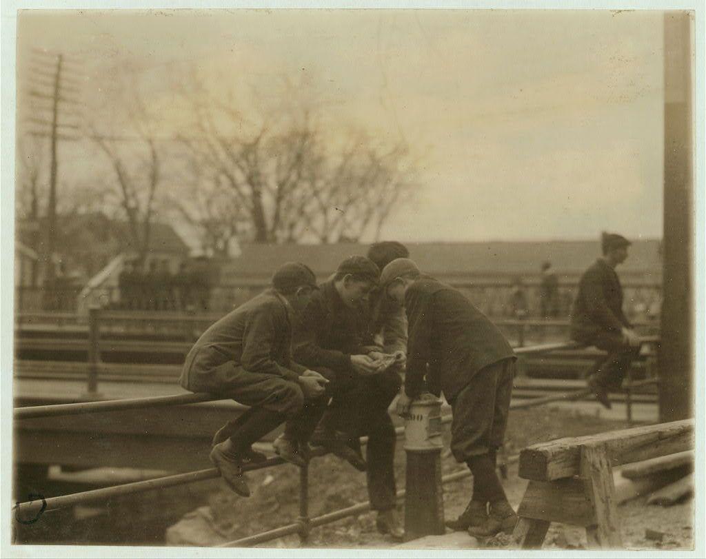 The image depicts a sepia-toned photograph capturing four individuals identified as "Mill Boys" in Lawrence, Massachusetts. These young men are clad in early 20th-century work attire, including dark suits and hats, indicative of the era's industrial labor fashion. They're seated on what appears to be wooden benches or platforms along an elevated railway track.

The setting is outdoors with overcast skies, suggesting a bleak working environment typical for factory workers during that period. Bare trees in the background imply it might be late fall or winter. The boys seem engaged in casual conversation and leisurely activities rather than work-related duties; two are leaning on metal bars of what could be part of railway infrastructure.

The composition includes another figure, possibly a supervisor or older worker, standing slightly apart from them, observing their interactions with the game at hand. This image is historically significant as it reflects working conditions for young males in textile mills during Lewis Hine's documented period.

Further contextual information notes that this photograph might be part of Noon-hour card games captured by Lewis Hine to depict social and labor environments where children were employed in early American industrial settings, such as the Pacific Mills.