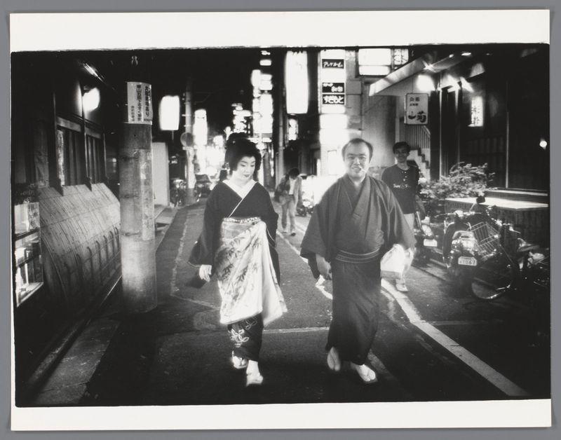 The image depicts a man and woman walking down an urban street at night. The man is dressed in traditional Japanese attire, consisting of a dark robe known as a yukata with sandals on his feet. He appears to be middle-aged or older. Beside him walks a younger-looking individual wearing what seems like a kimono, paired with lighter-toned pants and slippers similar to the woman's footwear. The street is illuminated by various signs and lights from storefronts, suggesting an urban setting possibly in Japan given their traditional garments.

The photo captures both figures mid-stride, conveying movement as they traverse the pavement together. Behind them, a parked bicycle and another pedestrian can be seen further down the road, adding to the sense of daily life in this cityscape scene. The image is rendered in black and white, emphasizing contrasts between light and shadow across its surface.

This particular photograph was taken by Ed van der Elsken, as indicated from archival records or associated information provided. Van der Elsken's work often captures street scenes with a candid yet thoughtful eye for composition and mood, reflecting an era when such snapshots of urban life were becoming increasingly significant in documenting cultural shifts and everyday experiences.

The timestamp within the image suggests it was taken between 1980 to 1990. The choice of clothing wor [...]