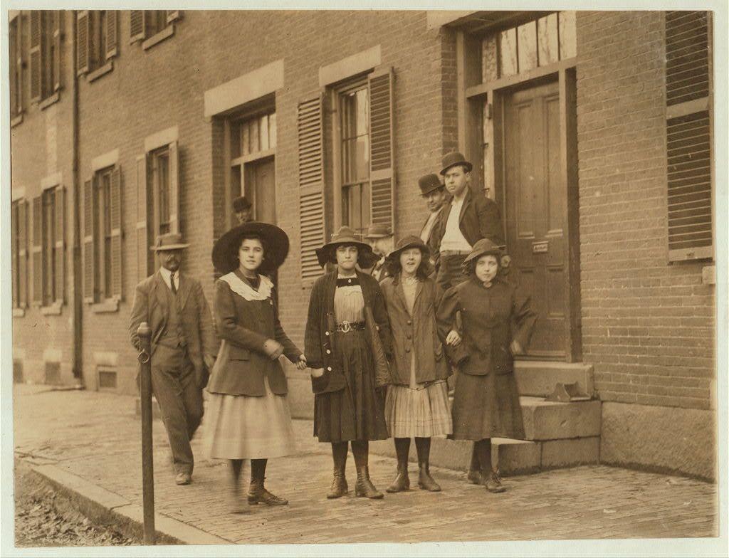The photo shows a group of seven people standing on the sidewalk outside brick buildings with windows and doors, possibly part of an industrial complex. There are five women dressed in early 20th-century fashion: one wearing a wide-brimmed hat and coat, three others holding hands; each wears dresses or coats suitable for work wear, likely indicating they could be workers at Pacific Mills mentioned in the caption text. Two men stand behind them, also attired formally but without hats; their presence suggests supervision roles.

Their attire is conservative with knee-length skirts on women and long pants on men, paired with dark shoes or boots. The overall mood appears somber due to the grayscale image quality of sepia tones common in early 20th-century photographs used for documentation rather than artistic expression.

The setting indicates a work environment possibly connected to Lawrence's textile industry during Lewis Hine’s period where he was known to document child labor conditions, reflecting on industrial life and social issues. The caption "Mill girls" refers specifically to the women depicted as part of such an industry workforce.