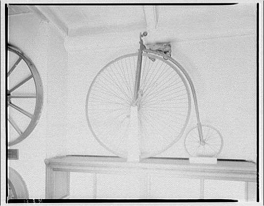 The image displays an old-fashioned bicycle suspended from the ceiling of a room with plain, light-colored walls. The bike has two large wheels connected by a straight frame without any pedals or seat; it features spoked rims and is positioned against what appears to be part of another wheel mechanism on the left side.

In addition to the vintage-style bicycle, there's also an old-fashioned wagon wheel visible in the background, mounted horizontally along with some other indistinct items. The setting suggests a historical context, possibly within a museum or educational institution showcasing antique transportation artifacts from around 1920-1950 era as mentioned by Horydczak.

The photograph is black and white which adds to its vintage feel. There's no discernible human presence in the image; only the bicycle serves as the focal point, highlighting a piece of history likely associated with Smithsonian Institution interiors according to accompanying information from an external source linked within this context[^1^].