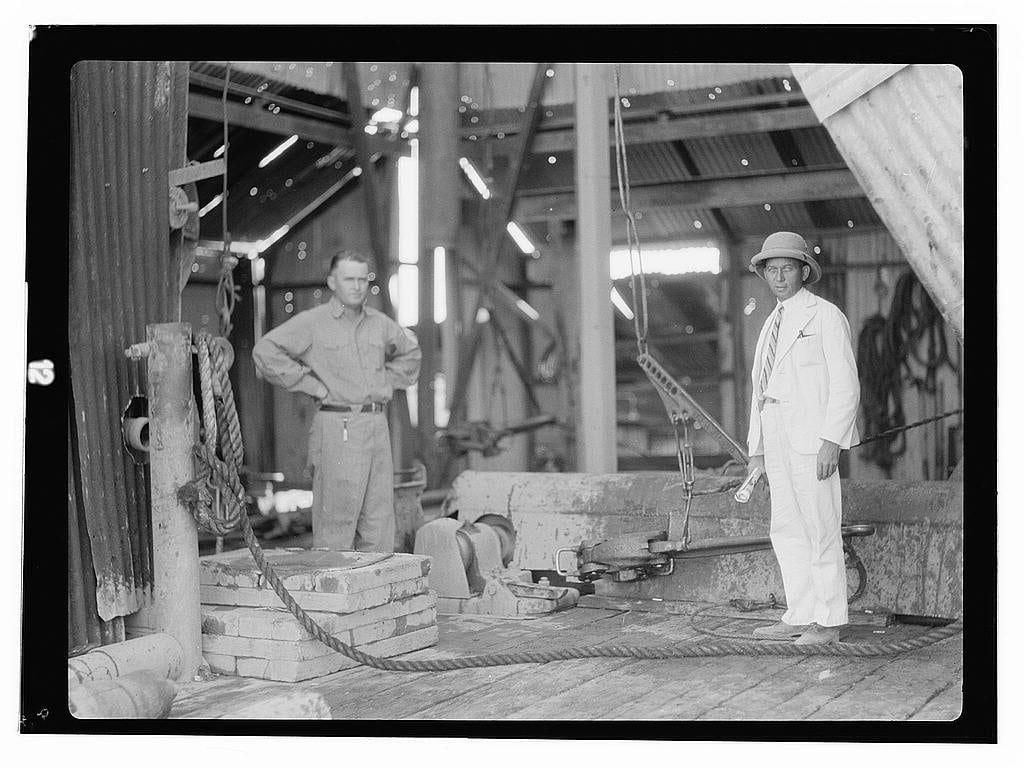 The image depicts a scene from an oil drilling operation, likely in the early 20th century. There are two men visible; one dressed in heavy-duty work attire with his hands on his hips and legs apart for stability, while another stands confidently to the right wearing light-colored clothing that looks like it could be part of a uniform or formal dress code.
The setting is an industrial environment featuring metal beams, ropes, barrels, and other equipment. The grayscale nature suggests this photo was taken in historical context when black-and-white photography dominated. There's no direct evidence indicating specific locations beyond the general mention of Iraq Petroleum Company near Kirkuk District. The image has a documentary feel to it, likely sourced from an archive related to American activities or interests during that period.
The men seem focused and engaged with their surroundings, which could imply a professional atmosphere at work sites like oil derricks in 1932.