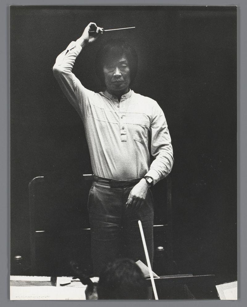 The image depicts a man standing on what appears to be an elevated platform or stage. He is holding something with his right hand, possibly a baton, which suggests he may be conducting an orchestra or leading some form of performance. The individual has dark hair and wears casual attire consisting of a light-colored long-sleeve shirt tucked into trousers, paired with shoes that appear to have laces. His stance exudes confidence as if in the middle of delivering instructions or expressing his interpretation during a musical piece. Behind him is darkness, possibly indicating an indoor setting such as a concert hall or theater. The photo captures this moment from a lower angle, focusing on the man's upper body and part of what seems to be sheet music at the bottom right corner near another person who may also be involved in the performance but not fully visible within the frame.