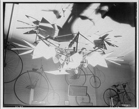 The image depicts an intriguing collection of antique bicycles displayed within a room adorned with overhead geometric decorations, possibly resembling pinwheels or origami. The black and white photograph captures the vintage charm of early bicycle models hanging in various orientations on the walls while some are placed near each other on stands. There's also visible text that reads "Bicycles at Smithsonian Institution ca. 1920-ca. 1950" suggesting a historical context for this exhibit, likely from around two to thirty years prior or after mentioned dates.

The photograph is credited to Theodor Horydczak (approximately born in the late 1800s and died early-mid twentieth century) who took one negative on safety film measuring eight by ten inches.