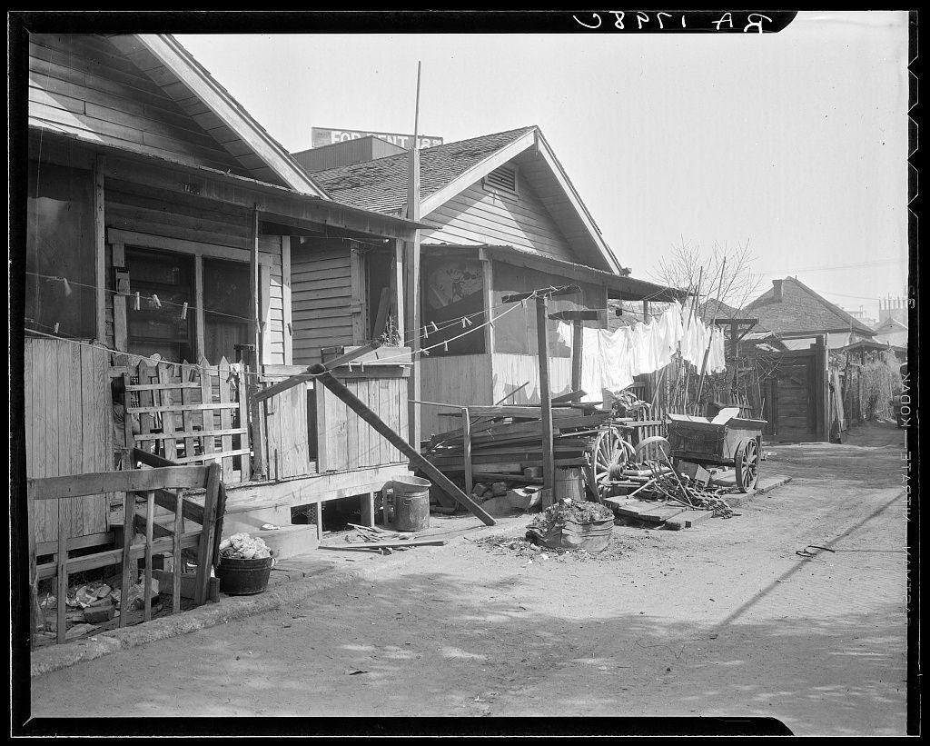 This black and white photograph depicts a rustic street scene, likely taken during the early 20th century. The main focus is on an old wooden house with a steeply pitched roof that has several items hung from its eaves: clotheslines stretched out to dry laundry against one wall; various household goods such as pots and pans displayed outside in front of windows. In close proximity, another structure houses more drying linens. An assortment of farming implements—like plows and cart wheels—are scattered around the area indicating a possible rural setting or agricultural activity.

The street is unpaved with visible dirt patches and debris that suggest wear from frequent use. A fence made of wood slats runs along one side while on another, there's what appears to be wooden pallets stacked together possibly used for storage or transport purposes. Various buckets, tools and a barrel rest against the buildings' exterior walls.

The architecture is indicative of modest residential structures with no visible modern amenities such as electricity meters indicating an era where utilities like gas lamps may have been more common than electric lights in homes during this period. The general atmosphere projects simplicity yet it reflects on how people lived then, possibly hinting at a working-class neighborhood or a community within Los Angeles known for its Mexican quarter and afforda [...]