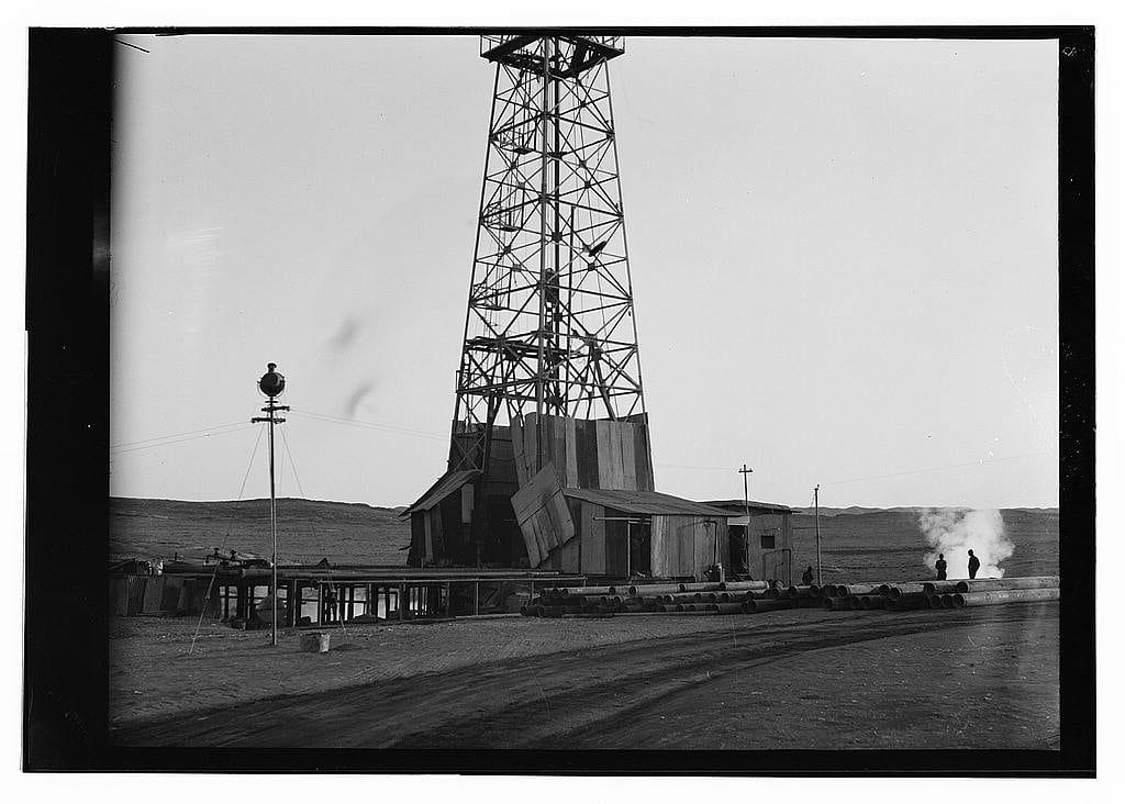The image is a black and white photograph depicting an oil drilling tower, possibly situated in the Iraq Petroleum Company's Kirkuk district. The towering structure stands prominently against a plain sky with no visible clouds or birds.
A wooden shack-like building sits adjacent to the platform under the rig, likely serving as housing for workers during extended operations on-site. A few individuals can be seen at various distances from this central oil extraction setup: one appears near the top of the drilling tower possibly operating some equipment; another is standing by a structure with pipes running horizontally across it.
In front of these structures lies an open space, paved and marked presumably for operational vehicles or perhaps as pathways. A few men are scattered in this area, implying active site workmanship. In the foreground, we see the edge of what seems to be a road leading away from the scene into the distance under clear skies with no visible vegetation.
The environment appears arid and desolate, lacking any significant trees or greenery that might suggest life besides the human activity in this industrial setting. The overall ambiance is quite stark and barren, which complements the black-and-white aesthetic of the photograph and adds to its historical feel.