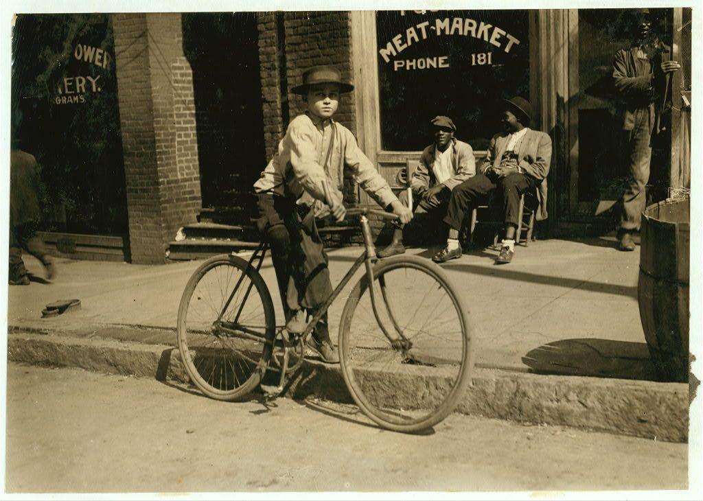 This black and white photograph captures a moment in the early 1900s, depicting an eleven-year-old delivery boy for an Opelika meat market. He is posed on his bicycle outside of the establishment, with three other individuals visible behind him. The child wears a hat and has a determined expression as he looks directly at the camera. His attire includes a light-colored jacket, trousers rolled up to reveal shoes, and possibly stockings or socks tucked into boots. The setting appears urban, likely in front of a meat market store indicated by signage on the building that reads "MEAT MARKET" along with additional information such as a phone number, suggesting this was an era before modern communication methods like cell phones became commonplace for small businesses to advertise their services through public directories or signs displayed prominently at storefronts. The presence of three other individuals indicates social interaction and possibly waiting customers outside the market during regular business hours in Opelika, Alabama; though specific times are not evident from this single snapshot captured within a historic image collection accessible via external link provided as reference material for further information on Lewis Hine's work or related historical context.