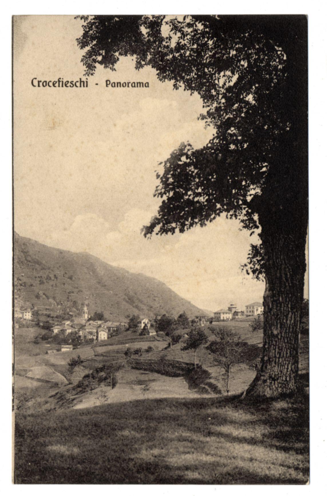 The image shows a sepia-toned panoramic photograph of the village of Crocefieschi. It features an expansive view with rolling hills in the background and buildings spread across the landscape, including what appears to be residential houses and possibly some larger structures that could serve as public or institutional purposes. The foreground is dominated by a large tree trunk on the right side, partially obscuring parts of the village scene behind it. The sky above has a cloudy appearance with soft edges, suggesting an overcast day. There's text at the top left corner indicating "Crocefieschi - Panorama," which likely refers to both the location and the type of image presented.